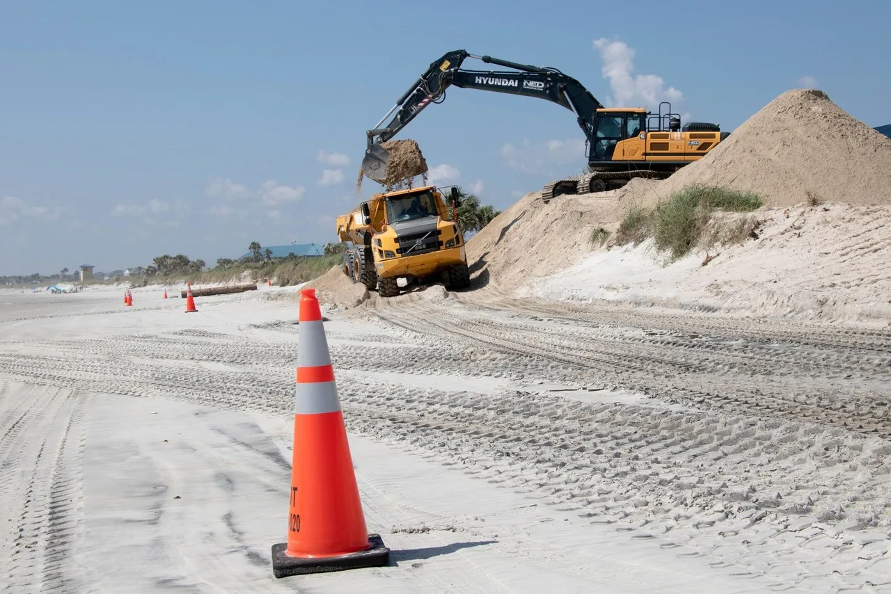 A Hyundai excavator on a sand dune loads a yellow articulated dump truck on the beach at Naval Station Mayport, Florida.