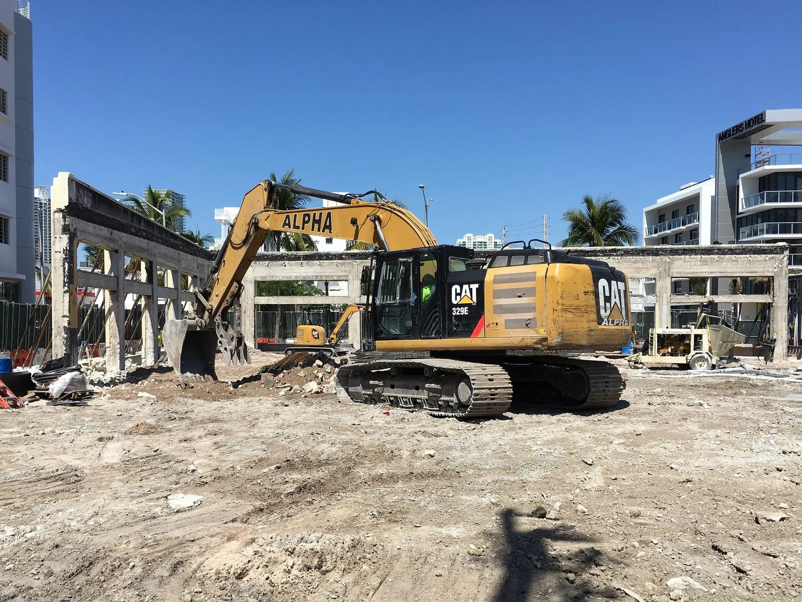 A yellow Caterpillar 329E excavator works a cleared lot in Miami Beach, Florida, under a deep blue sky, with palm trees and hotel towers behind it.